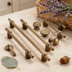 May include: An assortment of antique brass cabinet hardware, including bar pulls, knobs, and a keyhole pull. The hardware has a warm, aged finish. A basket of lavender and an acorn are in the background.