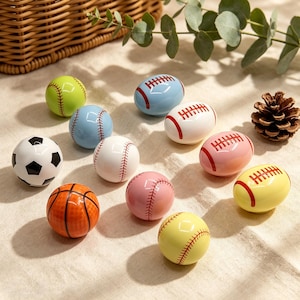 Peut inclure: Diverses sphères décoratives aux couleurs vives, représentant des ballons de sport. On y trouve des balles de baseball, de football américain, de basket-ball et de football. Les balles de baseball sont de différentes couleurs, avec des coutures rouges.