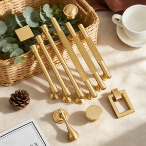 May include: A collection of gold-toned cabinet hardware, including handles, knobs, and a pull. The handles have a textured design, and the knobs are round. The hardware is arranged on a light-colored surface with a wicker basket and greenery in the background.