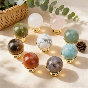 May include: A collection of nine gemstone spheres in various colors, including white, pink, green, brown, red, and blue, each mounted on a gold-colored base. A small pinecone and eucalyptus sprigs are also present.