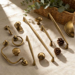 May include: An assortment of antique brass cabinet hardware, including knobs, pulls, and handles. The hardware features various designs, from simple round knobs to ornate pulls with curved details. Two small pinecones are also present.