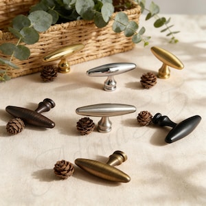 May include: A collection of cabinet pulls in various finishes, including gold, silver, bronze, and black. Each pull has a T-shaped design and is displayed with small pinecones. The pulls are arranged on a light-colored surface.