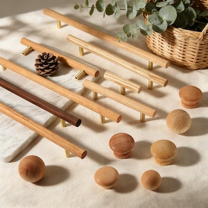 May include: A collection of wooden cabinet hardware, including various sizes of pulls and knobs. The pulls are cylindrical with brass-colored posts. The knobs are round and mushroom-shaped, showcasing natural wood grain. A pinecone and a wicker basket with greenery are also present.