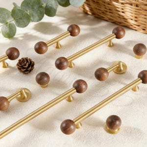 以下が含まれることがあります： 真鍮製の取っ手とノブを備えたキャビネットハードウェアのコレクション。濃い茶色の木製ボールのアクセントが付いています。取っ手は長く細く、ノブは丸いです。明るい色の表面に配置されています。