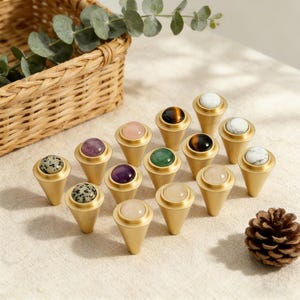 May include: A collection of decorative cabinet knobs with gold-colored bases and various gemstone tops, including amethyst, rose quartz, and tiger's eye. The knobs are arranged on a neutral surface, with a woven basket and greenery in the background.