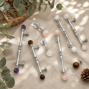 Può includere: Una collezione di maniglie e pomelli per mobili con finiture argentate lucide e dettagli in pietre preziose. Le maniglie presentano sfere di ametista, occhio di tigre, quarzo rosa e marmo bianco. Ferramenta decorativa per mobili.