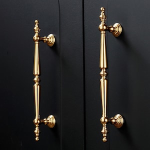 Op de afbeelding: Twee goudkleurige kastdeurgrepen tegen een zwarte achtergrond. De handgrepen zijn sierlijk, met een lange, slanke behuizing en decoratieve details aan de boven- en onderkant. De handgrepen zijn gemonteerd op een zwarte kastdeur.