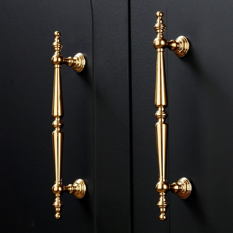 Op de afbeelding: Twee goudkleurige kastdeurgrepen tegen een zwarte achtergrond. De handgrepen zijn sierlijk, met een lange, slanke behuizing en decoratieve details aan de boven- en onderkant. De handgrepen zijn gemonteerd op een zwarte kastdeur.