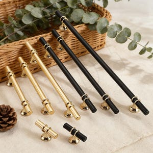 Puede incluir: Una variedad de tiradores y manijas de gabinete en dorado y negro. Los herrajes dorados incluyen tiradores de barra de varios tamaños y un pequeño pomo. Los herrajes negros incluyen tiradores de barra con detalles dorados. Todas las piezas tienen una base curva decorativa.