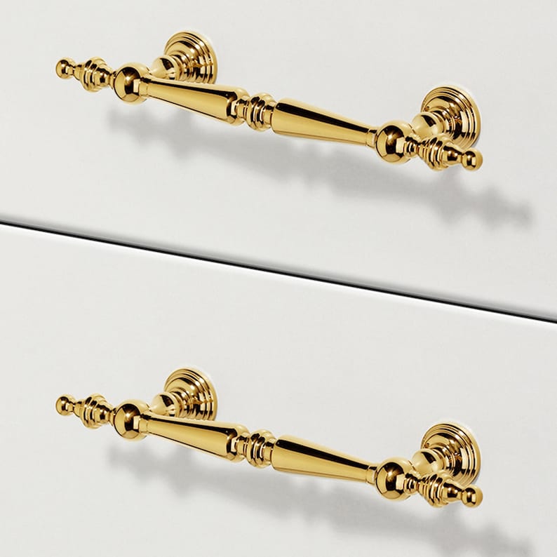 Op de afbeelding: Twee goudkleurige kastgrepen met sierlijke details. Elke greep heeft een centrale handgreep met decoratieve uiteinden en ronde montageplaten. De grepen zijn gemonteerd op een wit oppervlak, wat duidt op meubels of kasten.