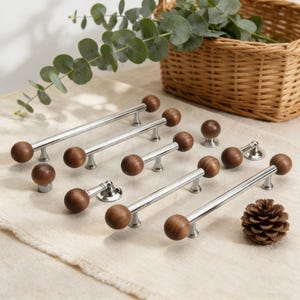 May include: A collection of cabinet hardware, including pulls and knobs. The pulls have a polished chrome finish with round, dark wood accents. The knobs are also dark wood. A pinecone and basket are in the background.