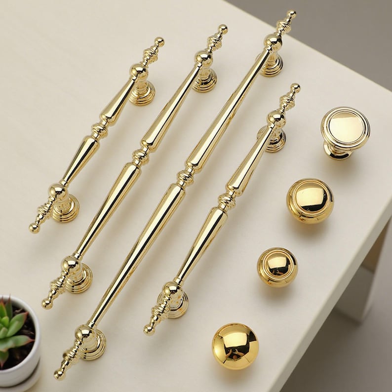 Op de afbeelding: Een verzameling goudkleurige kastbeslagen, waaronder vier lange handgrepen, twee ronde knoppen en drie bolvormige knoppen. De hardware heeft een glanzende afwerking en sierlijke details, geschikt voor meubels of kasten.