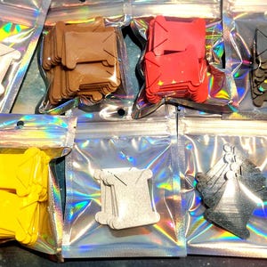 May include: Ten individual holographic resealable pouches, each containing a stack of plastic embroidery floss bobbins in various solid colors including pink, white, brown, red, black, lime green, yellow, grey, and dark red with star accents.