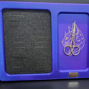 May include: A rectangular blue and purple organizer tray featuring a black foam insert with horizontal slots on the left side and a purple surface with a gold cat and moon constellation graphic on the right, holding small silver metal scissors.