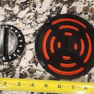 May include: Two black and orange circular objects are displayed next to a yellow measuring tape. The larger object has orange curved lines on a black background. The smaller object is black with white accents. The measuring tape shows inches.