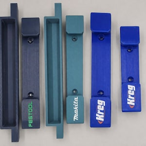 May include: Six vertical tool storage mounts in varying shades of blue and teal. The mounts feature brand logos including Festool, Makita, and Kreg. Each mount has a rectangular base with a protruding hook and pre-drilled holes for mounting.