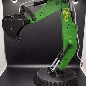 May include: A green and black toy excavator with a black bucket and base. The excavator arm has the John Deere logo. The toy is a miniature replica of a construction vehicle, suitable for play or display. The base is approximately 6 inches in diameter.