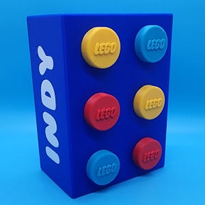 May include: A rectangular blue container featuring the white text INDY vertically on the side. The front surface displays six circular, raised LEGO-branded studs in yellow, red, and light blue, arranged in two columns of three, mimicking a building block.