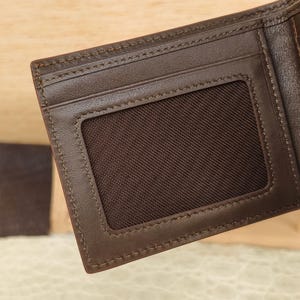 Puede incluir: Una cartera bifold de color marr&oacute;n oscuro que presenta una ranura rectangular para identificaci&oacute;n con una malla de color marr&oacute;n oscuro. La cartera est&aacute; construida con costuras tonales visibles a lo largo de los bordes de la ranura y el marco.