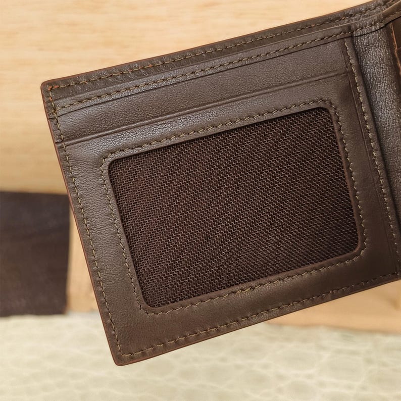 Puede incluir: Una cartera bifold de color marr&oacute;n oscuro que presenta una ranura rectangular para identificaci&oacute;n con una malla de color marr&oacute;n oscuro. La cartera est&aacute; construida con costuras tonales visibles a lo largo de los bordes de la ranura y el marco.