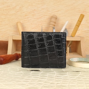 Puede incluir: Una cartera negra plegable con una textura brillante en relieve que imita la piel de cocodrilo. La cartera tiene forma rectangular con costuras visibles a lo largo de los bordes, mostrando una superficie oscura y reflectante.