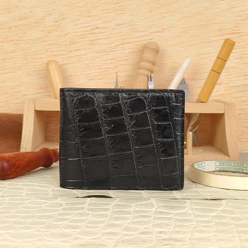 Puede incluir: Una cartera negra plegable con una textura brillante en relieve que imita la piel de cocodrilo. La cartera tiene forma rectangular con costuras visibles a lo largo de los bordes, mostrando una superficie oscura y reflectante.