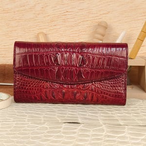 May include: A burgundy leather wallet with a crocodile skin texture. The wallet has a flap closure and a textured surface. The color is a deep, rich shade of red. The wallet is a stylish accessory.