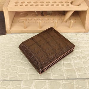 Puede incluir: Una cartera plegable de color marr&oacute;n oscuro con un patr&oacute;n de piel de cocodrilo brillante y texturizado. La cartera tiene forma rectangular con costuras visibles a lo largo de los bordes y un acabado liso y pulido que refleja la luz.