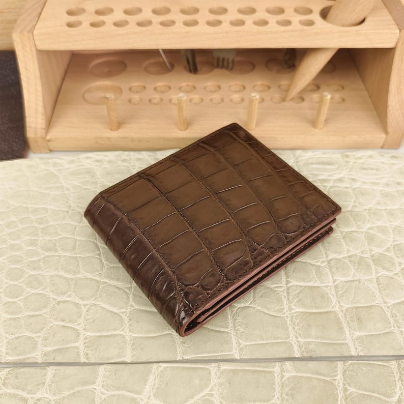 Puede incluir: Una cartera plegable de color marr&oacute;n oscuro con un patr&oacute;n de piel de cocodrilo brillante y texturizado. La cartera tiene forma rectangular con costuras visibles a lo largo de los bordes y un acabado liso y pulido que refleja la luz.