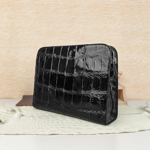 May include: A rectangular black clutch bag featuring a glossy finish and a prominent embossed crocodile skin texture. The bag has a structured silhouette with a silver-toned metal zipper closure along the top edge and a matching leather zipper pull.