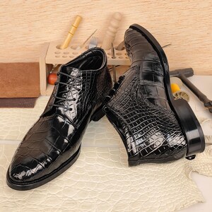 May include: A pair of black leather ankle boots with a crocodile skin texture. The boots have a lace-up design and a glossy finish. The shoes are displayed on a light-colored surface.