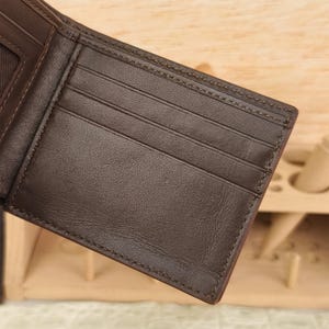 Puede incluir: Una cartera bifold de color marr&oacute;n oscuro que presenta m&uacute;ltiples ranuras horizontales para tarjetas en el panel interior. La cartera est&aacute; construida con costuras tonales visibles a lo largo de los bordes y compartimentos, con una textura mate.