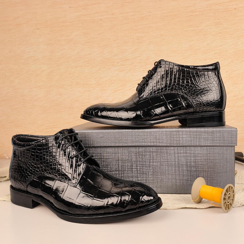 May include: Pair of black leather ankle boots with a crocodile skin texture. The boots are laced up and have a glossy finish. They are displayed on a gray box, suggesting a product presentation.