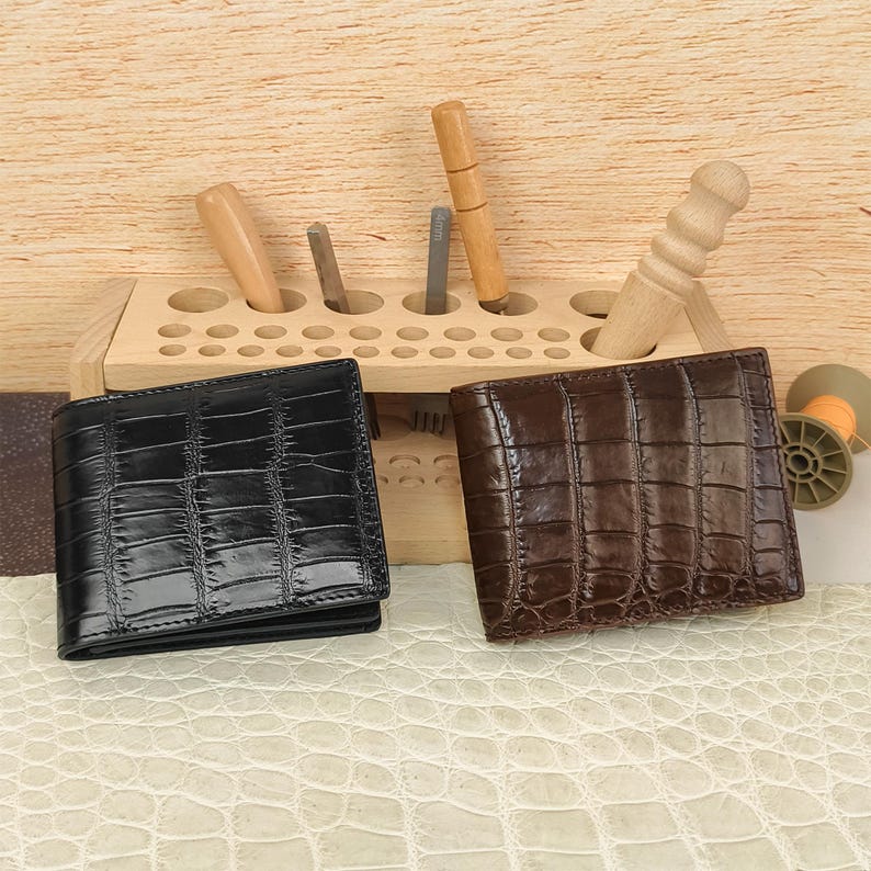 Puede incluir: Una cartera negra y una cartera marr&oacute;n oscuro, ambas con una textura brillante de piel de cocodrilo. Las carteras est&aacute;n colocadas frente a un organizador de herramientas de madera clara que contiene varias herramientas de metal y madera.