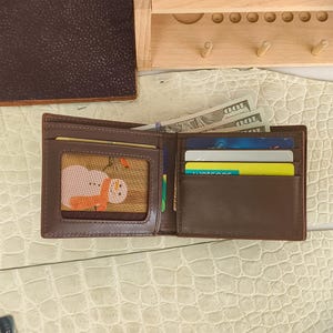 Puede incluir: Una cartera plegable de cuero marr&oacute;n con una ventana transparente que contiene un gr&aacute;fico de un mu&ntilde;eco de nieve, varias ranuras para tarjetas de cr&eacute;dito y un compartimento para billetes con billetes de cien d&oacute;lares.