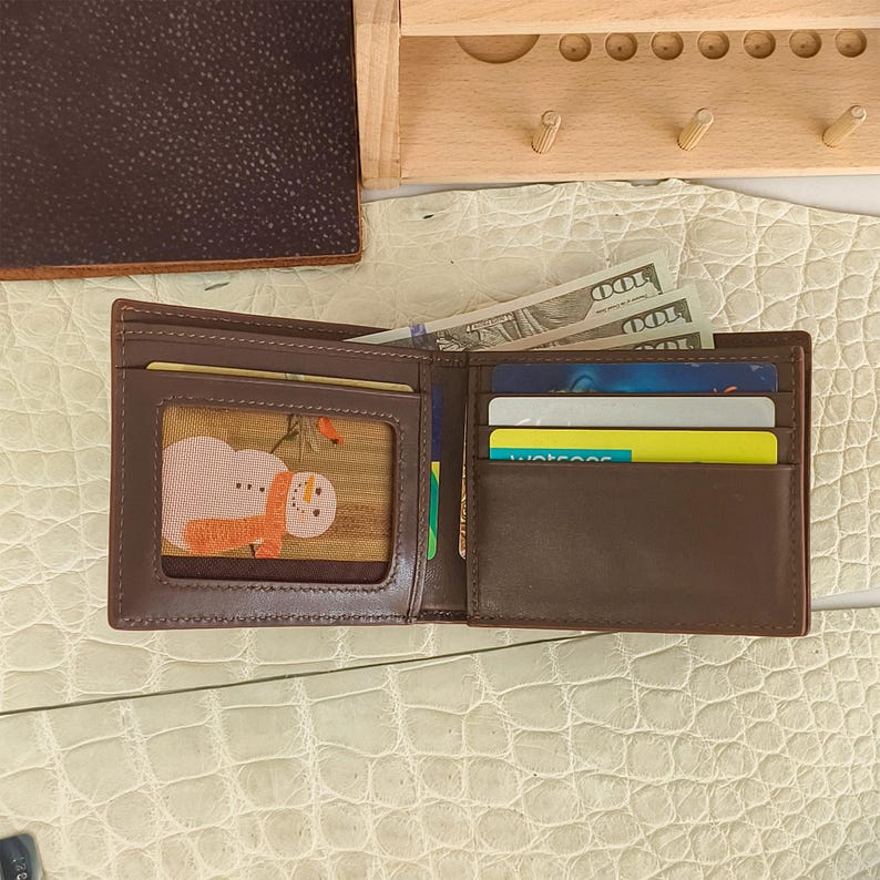 Puede incluir: Una cartera plegable de cuero marr&oacute;n con una ventana transparente que contiene un gr&aacute;fico de un mu&ntilde;eco de nieve, varias ranuras para tarjetas de cr&eacute;dito y un compartimento para billetes con billetes de cien d&oacute;lares.