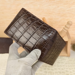 Puede incluir: Una cartera plegable de color marr&oacute;n oscuro con un patr&oacute;n de piel de cocodrilo brillante y texturizado. La superficie exterior muestra un dise&ntilde;o distintivo de escamas rectangulares repetitivas con un acabado pulido que resalta la veta natural.