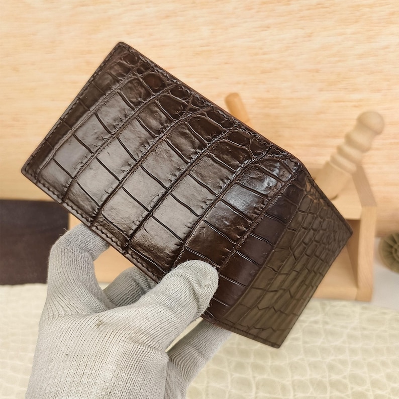 Puede incluir: Una cartera plegable de color marr&oacute;n oscuro con un patr&oacute;n de piel de cocodrilo brillante y texturizado. La superficie exterior muestra un dise&ntilde;o distintivo de escamas rectangulares repetitivas con un acabado pulido que resalta la veta natural.