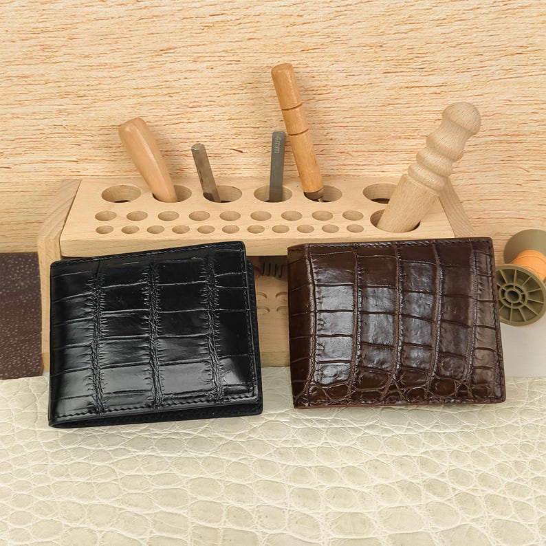 Puede incluir: Dos carteras plegables con una textura brillante de piel de cocodrilo. Una cartera es de color negro s&oacute;lido y la otra es de color marr&oacute;n oscuro. Est&aacute;n colocadas frente a un organizador de herramientas de madera clara con varios utensilios.