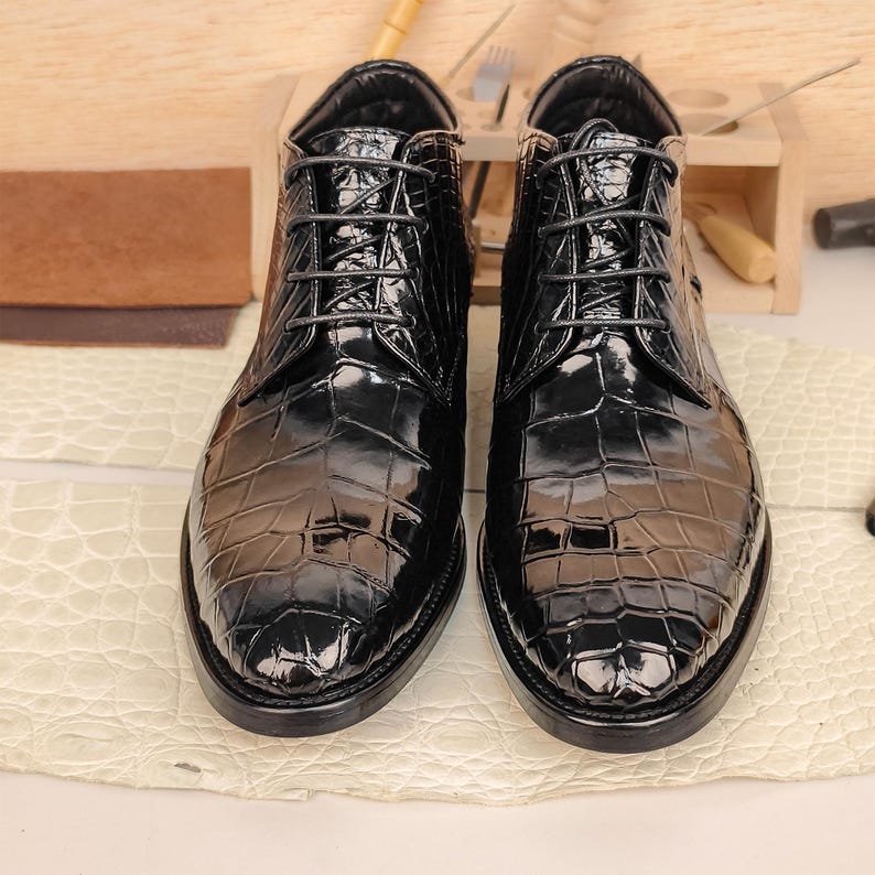 May include: Pair of glossy black leather ankle boots with a crocodile skin pattern. The boots have a lace-up design and a rounded toe. The shoes are displayed on a light-colored surface, with shoemaking tools in the background.