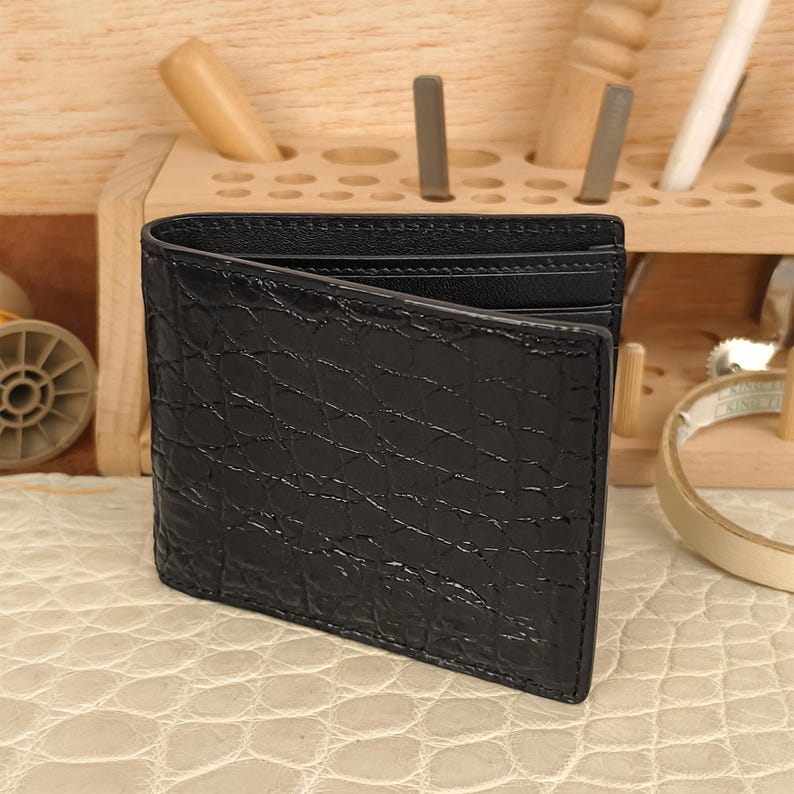 Puede incluir: Una cartera negra de dos pliegues con un patr&oacute;n de piel de cocodrilo texturizado en el exterior. La cartera est&aacute; construida con un forro interior negro liso e incluye m&uacute;ltiples ranuras para tarjetas visibles a lo largo del borde superior.