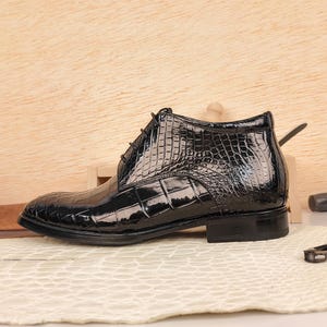 May include: A black, high-top dress shoe with a glossy, crocodile-skin texture. The shoe features a lace-up design and a low heel. The overall style is formal and elegant, suitable for special occasions or professional settings.