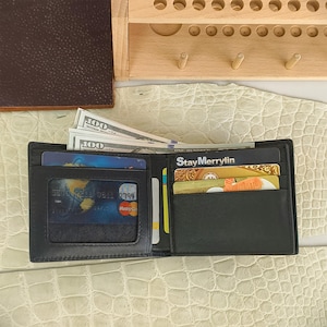 Puede incluir: Una cartera negra plegable con una ventana transparente para identificaci&oacute;n en el lado izquierdo que contiene una tarjeta con un gr&aacute;fico de globo terr&aacute;queo, y varias ranuras en el lado derecho con tarjetas, incluyendo una con StayMerrylin y billetes.