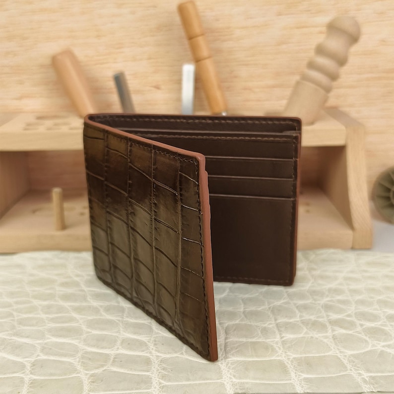 Puede incluir: Una cartera bifold de color marr&oacute;n oscuro con una textura de piel de cocodrilo brillante en el exterior. El interior est&aacute; forrado con cuero liso de color marr&oacute;n oscuro e incluye varias ranuras horizontales para tarjetas y un compartimento principal.