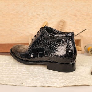 May include: A black, glossy leather boot with a crocodile skin pattern. The boot has a lace-up design and a low heel. The shoe is set on a cream-colored surface with a wooden background.