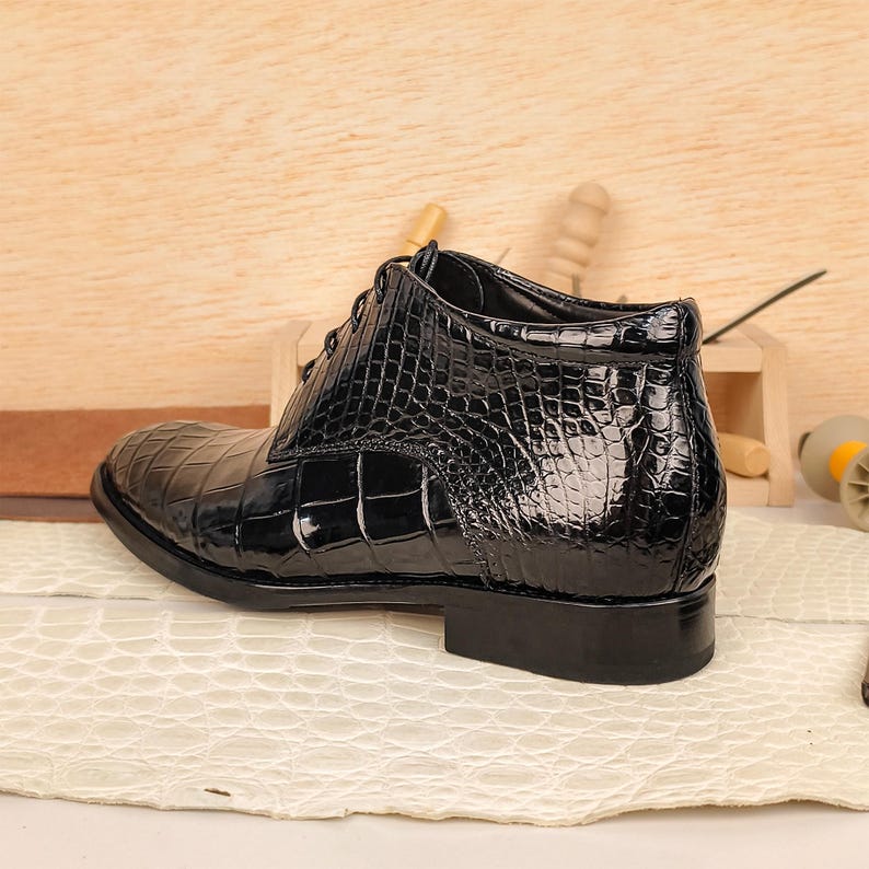 May include: A black, glossy leather boot with a crocodile skin pattern. The boot has a lace-up design and a low heel. The shoe is set on a cream-colored surface with a wooden background.