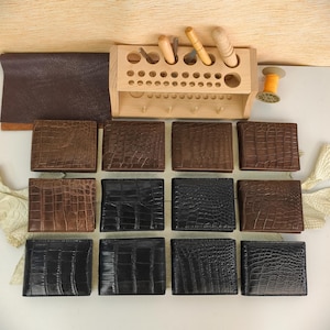 Puede incluir: Una colecci&oacute;n de doce carteras plegables dispuestas en tres filas, con cuero texturizado con relieve de cocodrilo en tonos negro y marr&oacute;n. Un organizador de herramientas de madera con varios utensilios de marroquiner&iacute;a y un carrete de hilo est&aacute;n arriba.
