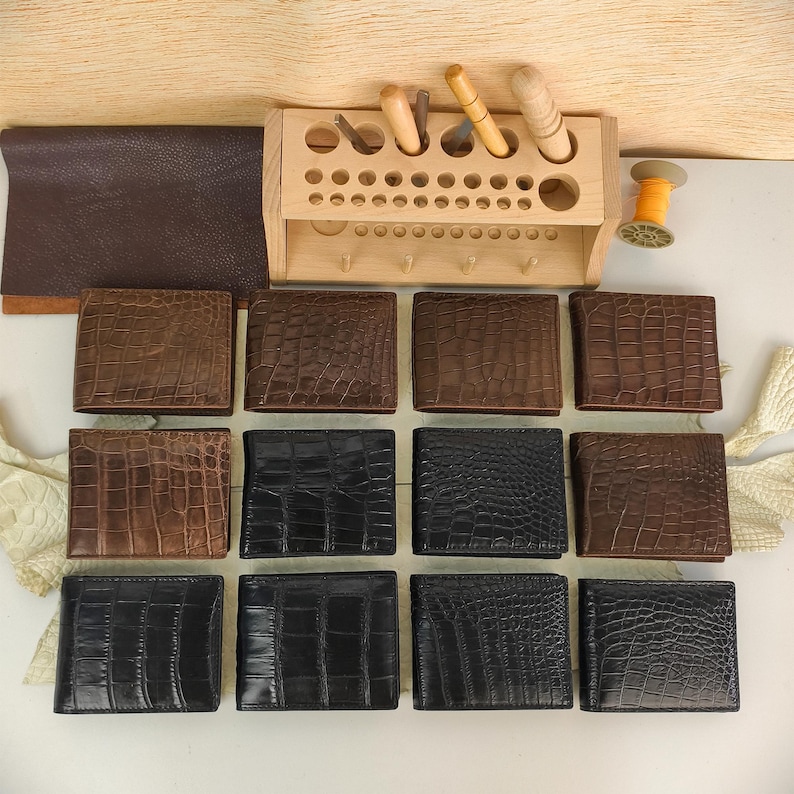 Puede incluir: Una colecci&oacute;n de doce carteras plegables dispuestas en tres filas, con cuero texturizado con relieve de cocodrilo en tonos negro y marr&oacute;n. Un organizador de herramientas de madera con varios utensilios de marroquiner&iacute;a y un carrete de hilo est&aacute;n arriba.