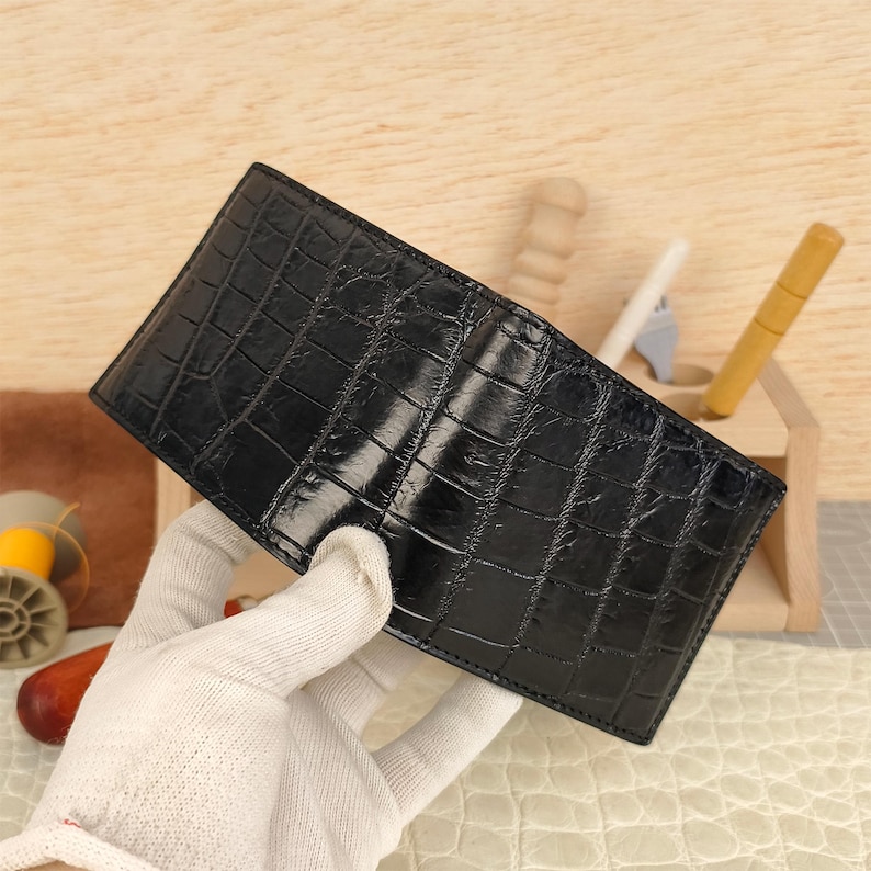 Puede incluir: Una cartera negra plegable con una textura brillante en relieve de piel de cocodrilo. El accesorio rectangular tiene un dise&ntilde;o estructurado y doblado con costuras visibles a lo largo de los bordes, mostrando un acabado oscuro y reflectante.
