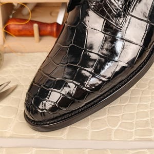 May include: Close-up of a black leather shoe with a glossy, crocodile-like texture. The shoe has a rounded toe and a dark sole. The leather reflects light, highlighting the texture and shape of the shoe.
